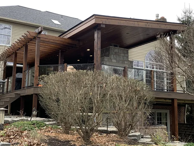 Covered deck and pergola with a stone fireplace and glass railing above a stone patio