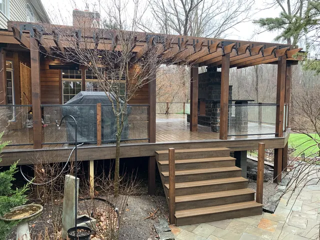 Raised deck with a pergola, glass railings, wide stairs and a stone fireplace